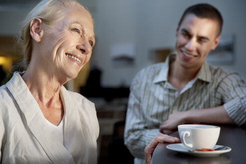 Lächelnde ältere Frau mit einer Tasse Kaffee und einem jungen Mann im Hintergrund - TOYF001364