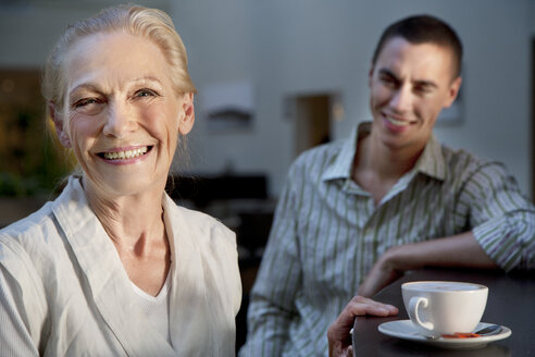 Porträt einer lächelnden älteren Frau mit einer Tasse Kaffee und einem jungen Mann im Hintergrund - TOYF001377