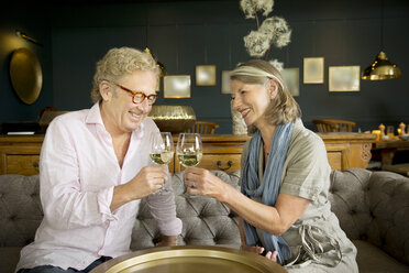 Lächelndes älteres Paar sitzt auf der Couch im Wohnzimmer und stößt mit Weingläsern an - TOYF001278