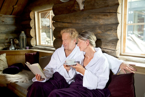 Älteres Paar sitzt in Bademänteln auf einer Bank, liest ein Buch und trinkt Tee - TOYF001286