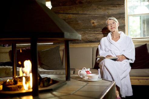 Ältere Frau sitzt im Bademantel auf einer Bank vor dem Kamin - TOYF001293