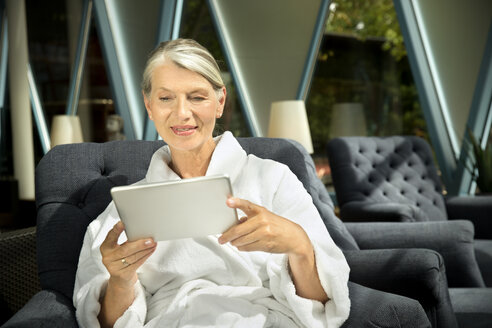 Ältere Frau im Bademantel sitzt im Sessel und benutzt ein digitales Tablet - TOYF001307
