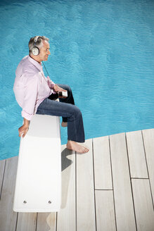 Spain, Mallorca, man sitting on springboard of a pool listening music with headphones - TOYF001176