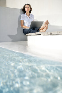 Spain, Mallorca, portrait of smiling woman sitting with laptop besides swimming pool - TOYF001177