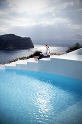 Spanien, Mallorca, Frau mit Laptop sitzt auf einer Treppe neben einem Schwimmbad mit dem Meer im Hintergrund - TOYF001179