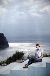 Spanien, Mallorca, Frau mit Laptop sitzt auf einer Treppe mit dem Meer im Hintergrund - TOYF001182