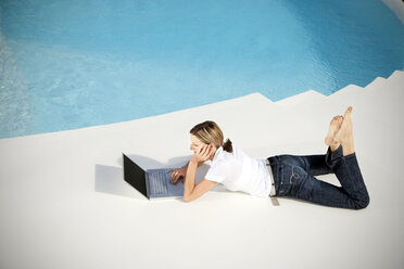 Spain, Mallorca, woman lying besides a swimming pool looking at laptop - TOYF001185