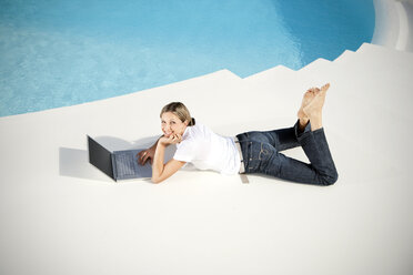 Spanien, Mallorca, lächelnde Frau mit Laptop neben einem Schwimmbad liegend - TOYF001186