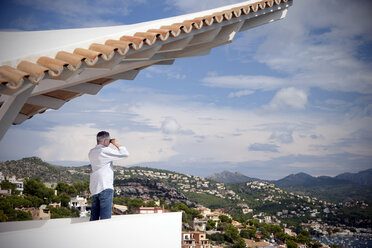 Spain, Mallorca, man standing on terrace of a house looking at view with binocular - TOYF001411