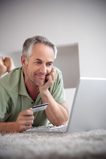 Portrait of smiling man lying with laptop on a carpet holding credit card - TOYF001201