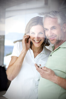 Portrait of smiling couple with earphones and mp3 player looking through window - TOYF001208