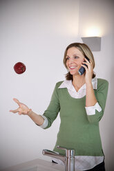 Portrait of smiling woman playing with an apple while telephoning in the kitchen - TOYF001212