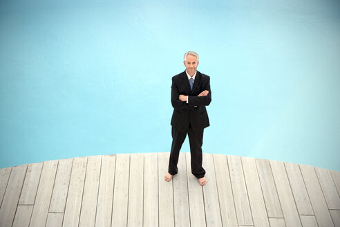 Barefoot businessman wearing black suit standing in front of swimming pool with arms crossed - TOYF001225