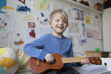 Portrait of smiling little boy sitting on his bed of his room playing guitar - RBF003000