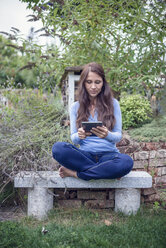 Frau sitzt im Schneidersitz auf einer Bank im Garten und schaut auf ein digitales Tablet - OPF000077