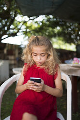 Little girl wearing red dress sitting on a chair using smartphone - RAEF000434