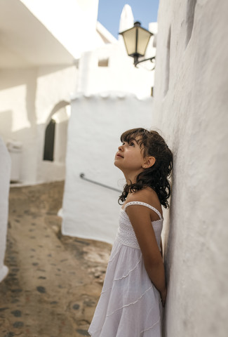 Spanien, Balearische Inseln, Menorca, Binibeca, kleines Mädchen lehnt an der Fassade und schaut nach oben, lizenzfreies Stockfoto