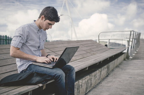 Spanien, Ferrol, junger Mann auf einer Bank sitzend mit Laptop - RAEF000488