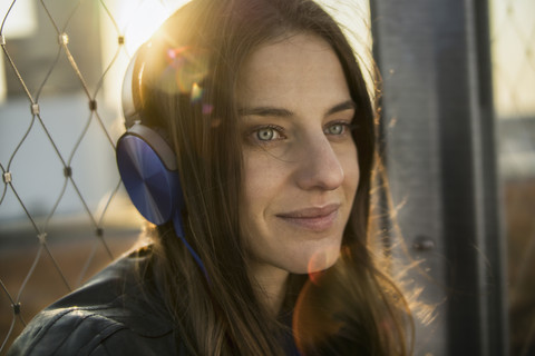 Deutschland, Frankfurt, Porträt einer lächelnden Frau, die mit Kopfhörern bei Gegenlicht Musik hört, lizenzfreies Stockfoto