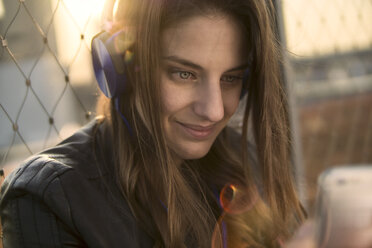 Germany, Frankfurt, portrait of smiling woman hearing music with headphones looking at smartphone - RIBF000258