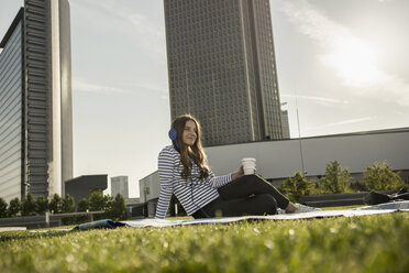 Deutschland, Frankfurt, junge Frau sitzt auf einer Decke auf einer Wiese und hört Musik mit Kopfhörern - RIBF000252