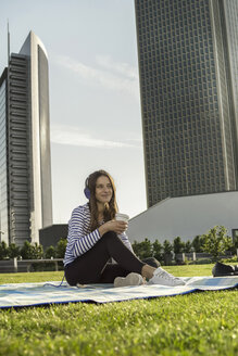 Deutschland, Frankfurt, junge Frau sitzt auf einer Decke auf einer Wiese und hört Musik mit Kopfhörern - RIBF000308