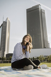 Deutschland, Frankfurt, junge Frau sitzt auf Decke auf einer Wiese und telefoniert mit Smartphone - RIBF000265