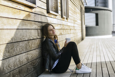 Lächelnde Frau mit Kaffee zum Mitnehmen sitzt auf dem Boden und lehnt sich an eine Holzwand - RIBF000269