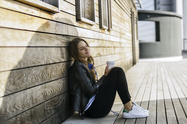 Smiling woman with coffee to go sitting on ground leaning against wooden wall - RIBF000269