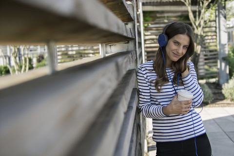 Porträt einer lächelnden Frau mit Kaffee zum Mitnehmen, die mit Kopfhörern Musik hört, lizenzfreies Stockfoto