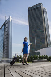 Germany, Frankfurt, businesswoman with smartphone and wheeled luggage standing on roof terrace - RIBF000298