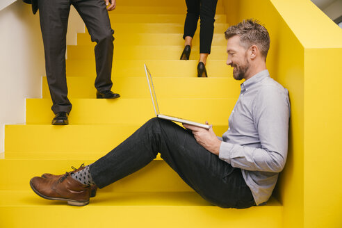 Smiling mature man sitting on yellow stairs with his laptop - MFF002123