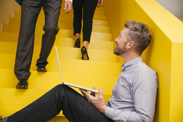 Mature man with laptop sitting on yellow stairs looking up to a colleague - MFF002124