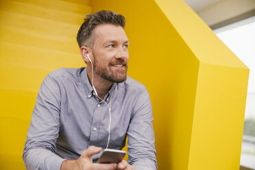 Portrait of smiling mature man hearing music with smartphone and earphones sitting on yellow stairs - MFF002126