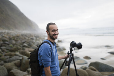 Spanien, Valdovino, Porträt eines lächelnden Fotografen am Strand mit Stativ und Kamera - RAEF000487