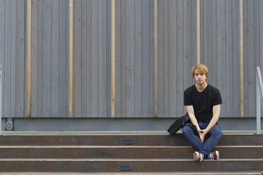 Junger Mann sitzt auf einer Treppe vor einer Holzfassade - SGF001873