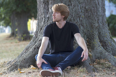 Pensive young man sitting in front of tree trunk - SGF001874