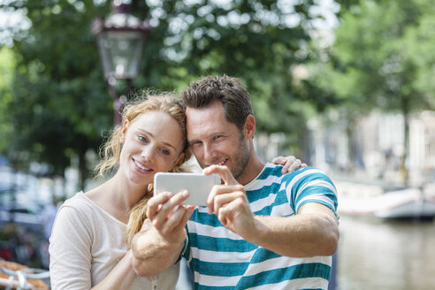 Niederlande, Amsterdam, lächelndes Paar, das ein Selfie am Stadtkanal macht - FMKF002094