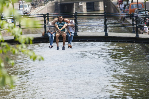 Niederlande, Amsterdam, drei glückliche Freunde sitzen auf einer Brücke am Stadtkanal - FMKF002098