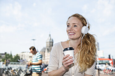 Niederlande, Amsterdam, lächelnde Frau mit Kopfhörern und Kaffee zum Mitnehmen in der Stadt - FMKF002109