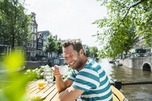 Niederlande, Amsterdam, glücklicher Mann trinkt eine Tasse Kaffee am Stadtkanal - FMKF002120