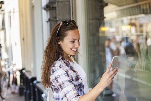 Niederlande, Amsterdam, lächelnde junge Frau in der Stadt, die auf ihr Mobiltelefon schaut - FMKF002135