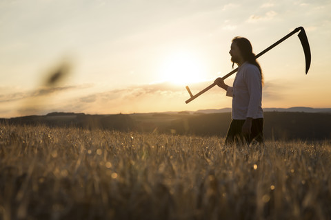 Biobauer mit Sense im Gerstenfeld bei Sonnenaufgang, lizenzfreies Stockfoto