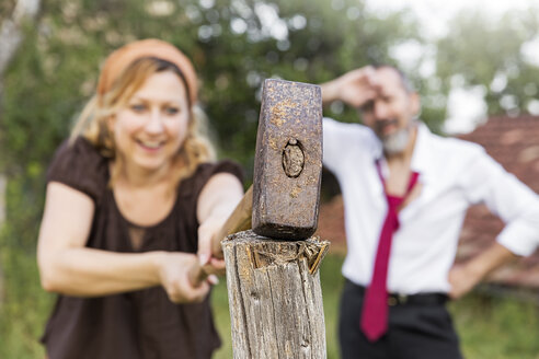 Woman hitting fence post with sledgehammer with exhausted businessman in the background - MIDF000616