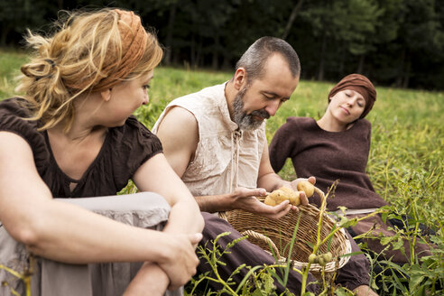 Three people sitting on field with potatoes in wicker basket - MIDF000626