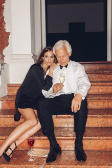Senior man and young woman with glasses of champagne on stairs - CHAF001445
