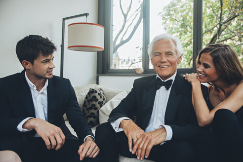 Smiling young woman with elegant senior man talking to young man on couch - CHAF001463