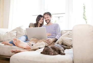 Smiling couple relaxing with laptop on the couch at living room - MFRF000394