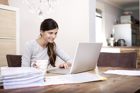 Lächelnde Frau sitzt mit einer Tasse Kaffee und einem Laptop an einem Holztisch - MFRF000419