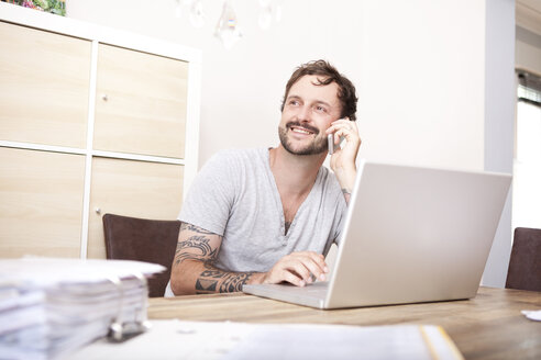 Lächelnder Mann sitzt an einem Holztisch mit Laptop und Ordner und telefoniert mit einem Smartphone - MFRF000425